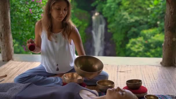 Superslowmotion filmet av en kvinnelig mester i asiatisk hellig medisin utfører tibetanske boller helbredende ritualer for en klient ung kvinne. Meditasjon med tibetanske sangboller. De er i et lysthus for... – stockvideo