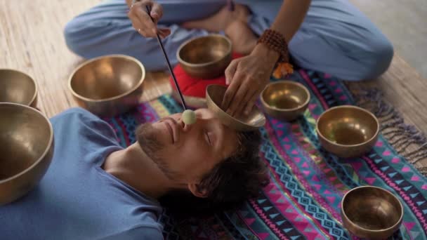 Superslowmotion tiro de uma mulher mestre da medicina sagrada asiática realiza tigelas tibetanas ritual de cura para um jovem cliente. Meditação com taças de canto tibetano. Eles estão em um gazebo para meditação — Vídeo de Stock