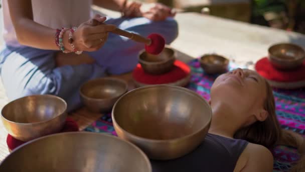 Superslowmotion skott av en kvinna mästare i asiatisk helig medicin utför tibetanska skålar helande ritual för en klient ung kvinna. Meditation med tibetanska sjungande skålar. De är i ett lusthus för — Stockvideo