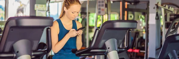 Молодая женщина пользуется телефоном во время тренировки в спортзале. Женщина сидит на тренажере с мобильным телефоном баннер, длинный формат — стоковое фото