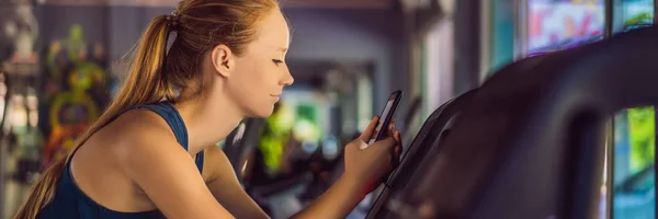 Jeune femme utilisant le téléphone pendant l'entraînement à la salle de gym. Femme assise sur une machine à exercer tenant un téléphone portable BANNER, LONG FORMAT — Photo