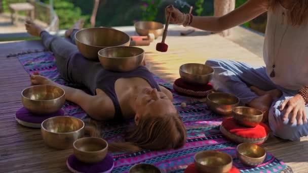 Superslowmotion skott av en kvinna mästare i asiatisk helig medicin utför tibetanska skålar helande ritual för en klient ung kvinna. Meditation med tibetanska sjungande skålar. De är i ett lusthus för — Stockvideo