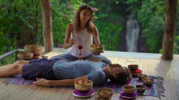 Superslowmotion shot of a woman master of Asian sacred medicine performs Tibetan bowls healing ritual for a client young man. Meditation with Tibetan singing bowls. They are in a gazebo for meditation — Stock Video
