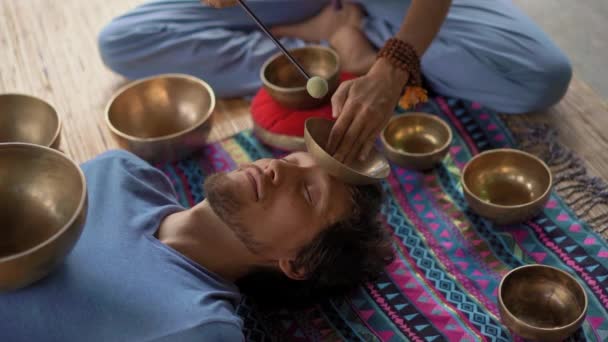Superslowmotion disparo de una mujer maestra de la medicina sagrada asiática realiza cuencos tibetanos ritual de curación para un cliente joven. Meditación con cuencos tibetanos. Están en un cenador para meditar. — Vídeo de stock