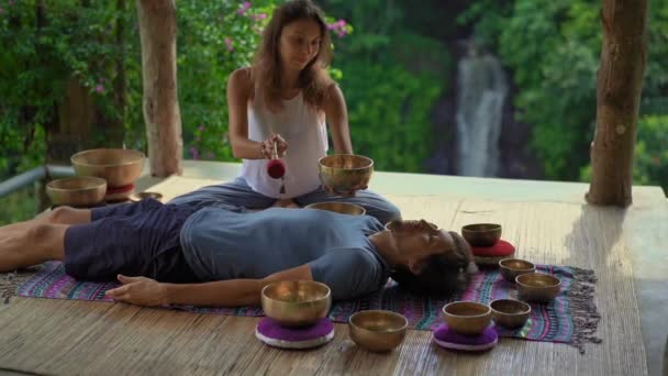 Superslowmotion shot of a woman master of Asian sacred medicine performs Tibetan bowls healing ritual for a client young man. Meditation with Tibetan singing bowls. They are in a gazebo for meditation — Stock Video