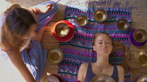 Superslowmotion tiro de uma mulher mestre da medicina sagrada asiática realiza tigelas tibetanas ritual de cura para uma jovem cliente. Meditação com taças de canto tibetano. Eles estão em um gazebo para — Vídeo de Stock