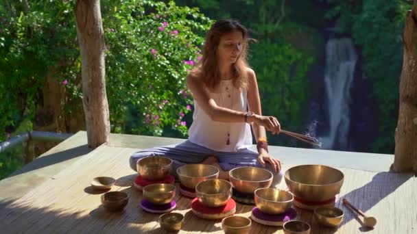 Superslowmotion tiro de uma mulher mestre da medicina sagrada asiática realiza tigelas tibetanas ritual de cura. Meditação com taças de canto tibetano. Ela se senta em um gazebo para meditação com uma bela — Vídeo de Stock