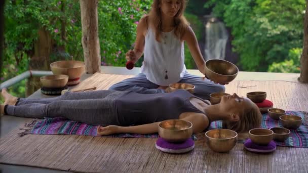 Superslowmotion disparo de una mujer maestra de la medicina sagrada asiática realiza cuencos tibetanos ritual de curación para una joven cliente. Meditación con cuencos tibetanos. Están en un mirador para — Vídeos de Stock