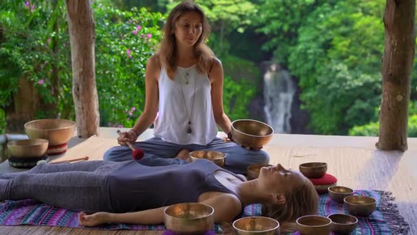 Superslowmotion shot of a woman master of Asian sacred medicine performs Tibetan bowls healing ritual for a client young woman. Meditation with Tibetan singing bowls. They are in a gazebo for — Stock Video