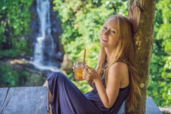 Closeup portrait image of a beautiful woman drinking ice tea with feeling happy in green nature and waterfall garden background — Stock Photo, Image