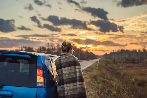 Una donna in un plaid si trova accanto alla macchina sul ciglio della strada sullo sfondo dell'alba. Concetto di viaggio — Foto Stock