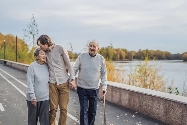 Een bejaarde echtpaar wandelingen in het park met een mannelijke assistent of volwassen kleinzoon. Zorg voor ouderen, vrijwilligerswerk — Stockfoto