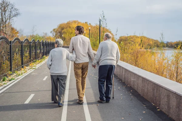 Ein älteres Ehepaar spaziert mit einem männlichen Assistenten oder erwachsenen Enkel durch den Park. Altenpflege, Freiwilligenarbeit — Stockfoto