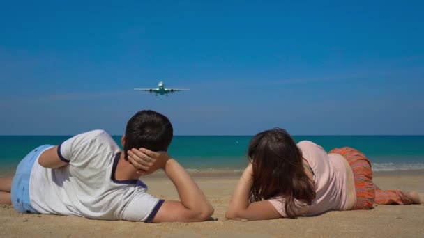 Junger Mann und Frau liegen an einem schönen Strand und beobachten ein landendes Flugzeug — Stockvideo