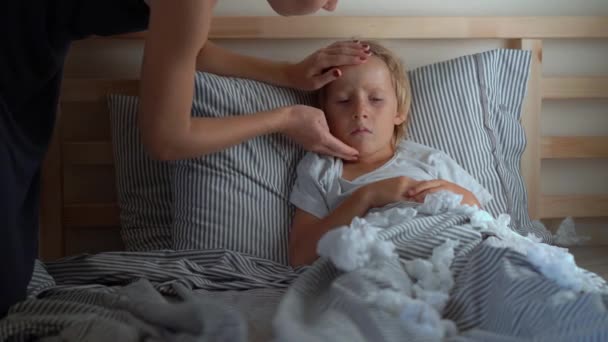 A sick little boy in a bed. Mother kisses him and measures his temperature. Baby flu concept — Stock Video