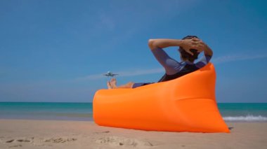 Tropik bir plajda şişme bir kanepede yatan mutlu turist superslowmotion çekim bir uçak iniş izlerken