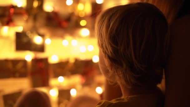 母親と彼女の小さな息子のスローモーションクローズアップショットは、クリスマスライトで明るいベッドにぶら下がっているアドベントカレンダーを見ます。クリスマスと新年のコンセプトの準備。アドベントカレンダー — ストック動画