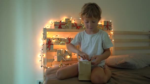 Zeitlupenaufnahme eines kleinen Jungen beim Öffnen eines Geschenks aus einem Adventskalender, das an einem mit Weihnachtslichtern beleuchteten Bett hängt. Wir bereiten uns auf Weihnachten und Neujahr vor. Advent