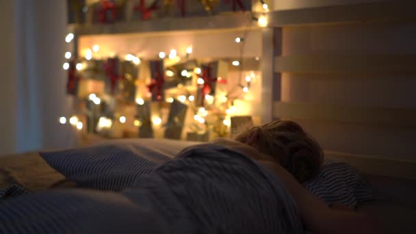 La toma en cámara lenta de un niño durmiendo en su cama con un calendario de advenimiento aligerado con luces de Navidad brilla en una parte posterior de su cama. Preparándose para la Navidad y el concepto de Año Nuevo. Calendario de Adviento — Vídeo de stock