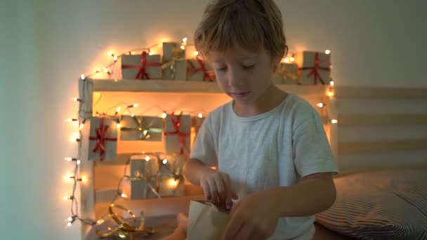 Slowmotion shot van een jongetje dat een geschenk opent vanuit een adventkalender die op een bed hangt dat verlicht is met kerstverlichting. Klaar voor het kerst-en Nieuwjaars concept. Komst — Stockvideo