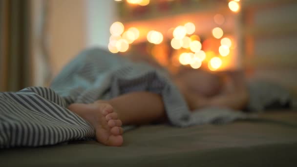 La toma en cámara lenta de un niño durmiendo en su cama con un calendario de advenimiento aligerado con luces de Navidad brilla en una parte posterior de su cama. Preparándose para la Navidad y el concepto de Año Nuevo. Calendario de Adviento — Vídeos de Stock