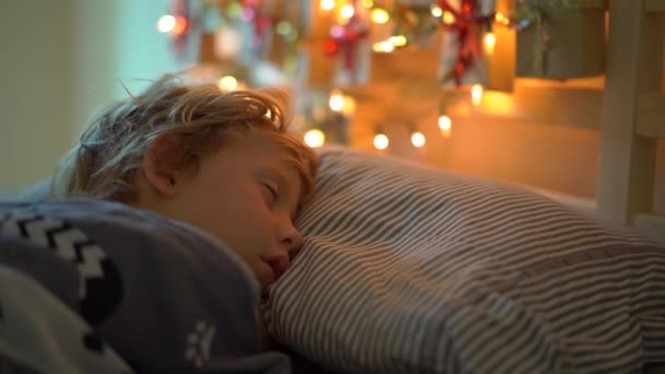 Slowmotion strzał małego chłopca śpi w łóżku z kalendarza adwentowego rozjaśnić z lampami świątecznymi świeci na tylnej części łóżka. Przygotowanie do Bożego Narodzenia i nowego roku koncepcji. Kalendarz adwentowy — Wideo stockowe