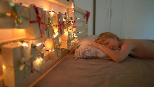 La toma en cámara lenta de un niño pequeño se despierta y ve un calendario de advenimiento colgado en una cama iluminada con luces de Navidad. Preparándose para la Navidad y el concepto de Año Nuevo. Concepto de calendario de Adviento — Vídeos de Stock