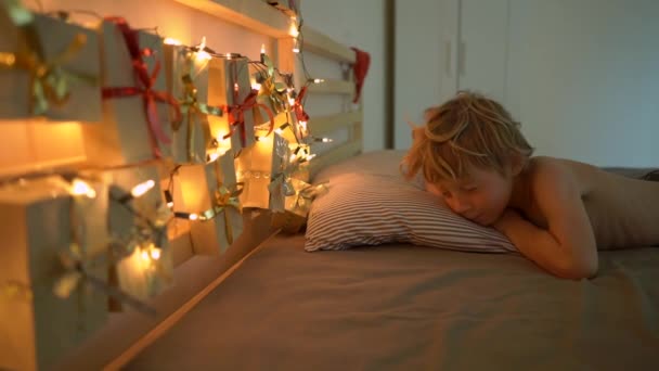 La toma en cámara lenta de un niño pequeño se despierta y ve un calendario de advenimiento colgado en una cama iluminada con luces de Navidad. Preparándose para la Navidad y el concepto de Año Nuevo. Concepto de calendario de Adviento — Vídeo de stock