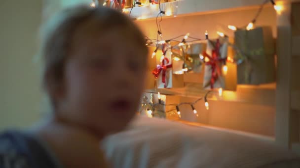 Slowmotion strzał małego chłopca budząc się w łóżku z kalendarza adwentowego rozjaśnić z lampami świątecznymi świeci na tylnej części łóżka. Przygotowanie do Bożego Narodzenia i nowego roku koncepcji. Adwentowy — Wideo stockowe