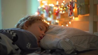 Slowmotion bir gelişme takvimi ile yatakta uyuyan küçük bir çocuk çekim Noel ışıkları ile hafifletmek onun yatağın bir arka parlar. Noel ve yeni yıl konsepti için hazırlanıyor. Advent takvimi