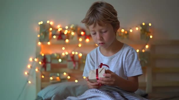 Tiro de câmera lenta de um menino abrindo um presente de um calendário de advento que está pendurado em uma cama que é iluminada com luzes de Natal. Preparando-se para o Natal e Ano Novo conceito. Advento — Vídeo de Stock