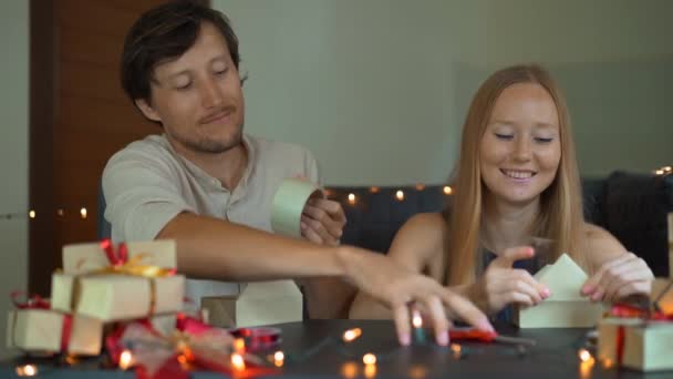 若い女性と男性の父と母のラッププレゼントのスローモーションショット。クリスマスや新年に赤と金のリボンでクラフトペーパーに包まれたプレゼント。親はアドベントカレンダーを作る — ストック動画