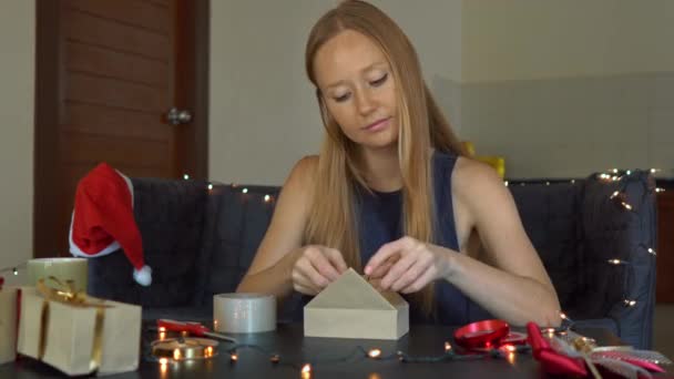 Eine junge Frau packt Geschenke ein. Geschenk eingewickelt in Bastelpapier mit einem rot-goldenen Band für Weihnachten oder Neujahr. Frau bastelt Adventskalender für ihre Kinder — Stockvideo