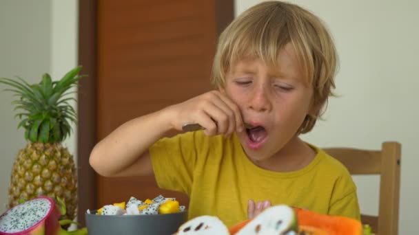 Sarı bir gömlek küçük bir çocuk bir tutku meyve veya Maracuja ve çok ekşi yiyor. Bir masa üzerinde döşeme tropikal meyve Lots onu çevreleyen. Sağlıklı gıda konsepti — Stok video