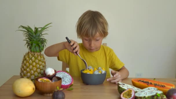 Um garotinho de camisa amarela come uma mistura de frutas de manga e dragão e muitas frutas tropicais deitadas em uma mesa o cercam. Conceito de alimentação saudável — Vídeo de Stock