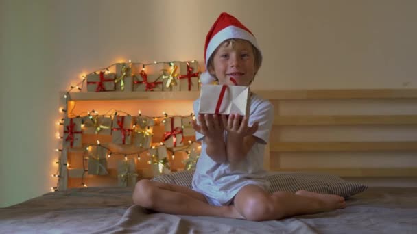 Een kleine jongen die een geschenk opent vanuit een adventkalender die op een bed hangt dat verlicht is met kerstverlichting. Klaar voor het kerst-en Nieuwjaars concept. Adventkalender concept — Stockvideo