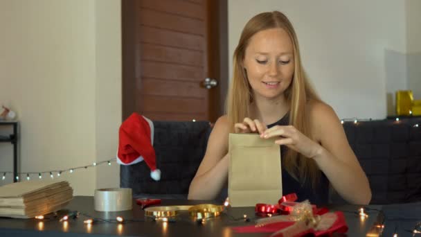 Una joven empacando regalos. Presente envuelto en papel artesanal con una cinta roja y dorada para Navidad o año nuevo. Mujer hace un calendario de adviento para sus hijos — Vídeo de stock