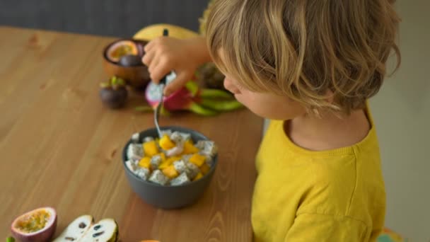 Sarı bir gömlek küçük bir çocuk mango ve ejderha meyvelerinin bir karışımı yiyor ve tropikal meyve bir masa üzerinde döşeme onu çevreleyen bir sürü. Sağlıklı gıda konsepti — Stok video