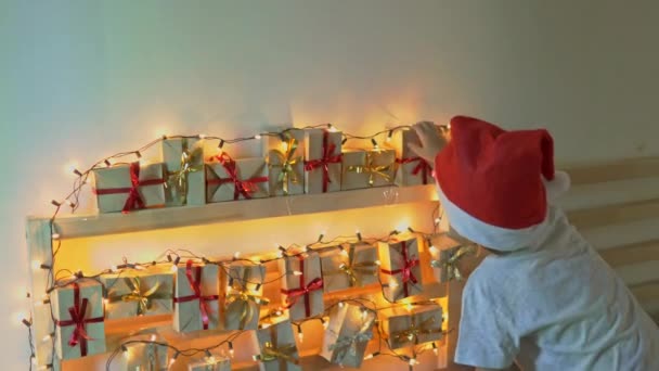Little boy takes a present from an advent calendar hanging on a bed that is lightened with Christmas lights. Getting ready for Christmas and New Year concept. Advent calendar concept — Stock Video