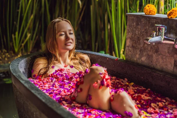 Atractiva Mujer joven en baño con pétalos de flores tropicales y aceites aromáticos. Tratamientos spa para el rejuvenecimiento de la piel. Mujer atractiva en el salón de Spa. Chica relajante en la bañera con pétalos de flores. Lujo — Foto de Stock