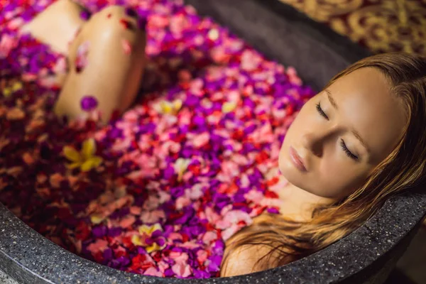 Atractiva Mujer joven en baño con pétalos de flores tropicales y aceites aromáticos. Tratamientos spa para el rejuvenecimiento de la piel. Mujer atractiva en el salón de Spa. Chica relajante en la bañera con pétalos de flores. Lujo — Foto de Stock