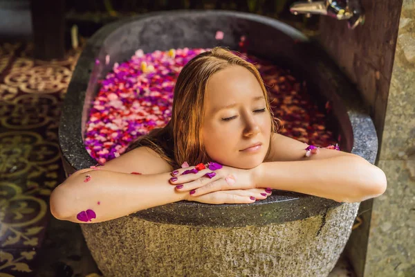 Atractiva Mujer joven en baño con pétalos de flores tropicales y aceites aromáticos. Tratamientos spa para el rejuvenecimiento de la piel. Mujer atractiva en el salón de Spa. Chica relajante en la bañera con pétalos de flores. Lujo — Foto de Stock