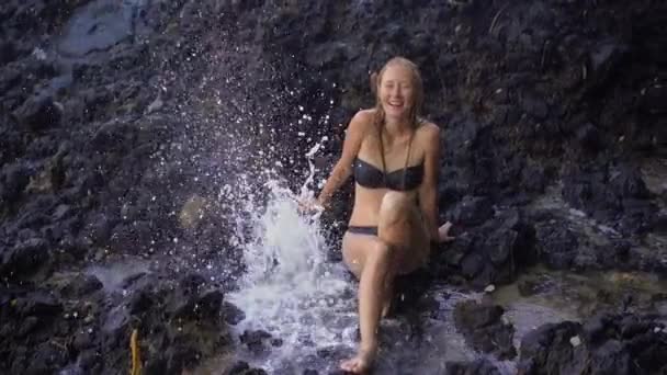 Slowmotion strzał młodej kobiety korzystających z fontanny lub gejzatory wykonane z natury na skały wulkaniczne w pobliżu białej piaszczystej plaży na wyspie Bali — Wideo stockowe