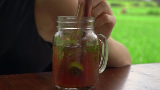 Syuting closeup seorang wanita muda minum es teh dengan mint dan lemon di kafe jalan dengan sawah di latar belakang — Stok Video