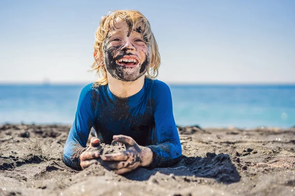 ブラックフライデーのコンセプト。海で泳ぐ前に、黒い砂浜に座って遊んで汚れた黒い顔をした笑顔の少年。家族のアクティブなライフスタイル、子供たちとの夏休みの水のレジャー — ストック写真