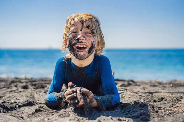 Black Friday concept. Lachende jongen met vuile zwarte gezicht zitten en spelen op zwarte zand zee strand voordat zwemmen in de Oceaan. Familie actieve levensstijl en watervrije tijd op zomer vakantie met kinderen — Stockfoto