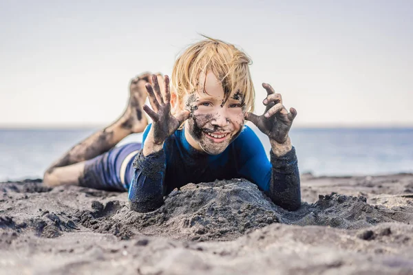 Black Friday concept. Lachende jongen met vuile zwarte gezicht zitten en spelen op zwarte zand zee strand voordat zwemmen in de Oceaan. Familie actieve levensstijl en watervrije tijd op zomer vakantie met kinderen — Stockfoto