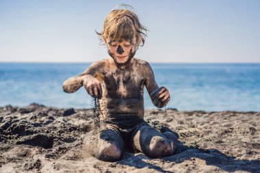 Kara Cuma konsepti. Okyanusta yüzmeden önce kirli Siyah yüzü oturan ve siyah kum denizi sahilinde oynayan gülümseyen çocuk. Aile aktif yaşam tarzı ve çocuklarla yaz tatilinde su eğlence