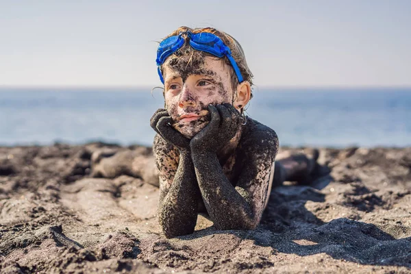 Koncepcja czarnego piątku. Uśmiechnięty chłopak z brudną czarną twarzą siedząc i grając na czarnym piasku morza plaży przed pływaniem w oceanie. Rodzinny aktywny styl życia i wodny wypoczynek na letnich wakacjach z dziećmi — Zdjęcie stockowe