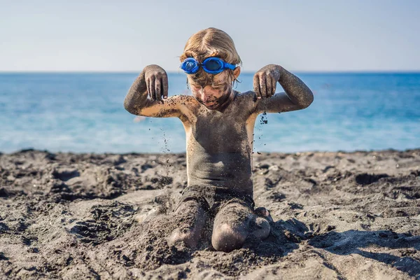 Koncepcja czarnego piątku. Uśmiechnięty chłopak z brudną czarną twarzą siedząc i grając na czarnym piasku morza plaży przed pływaniem w oceanie. Rodzinny aktywny styl życia i wodny wypoczynek na letnich wakacjach z dziećmi — Zdjęcie stockowe